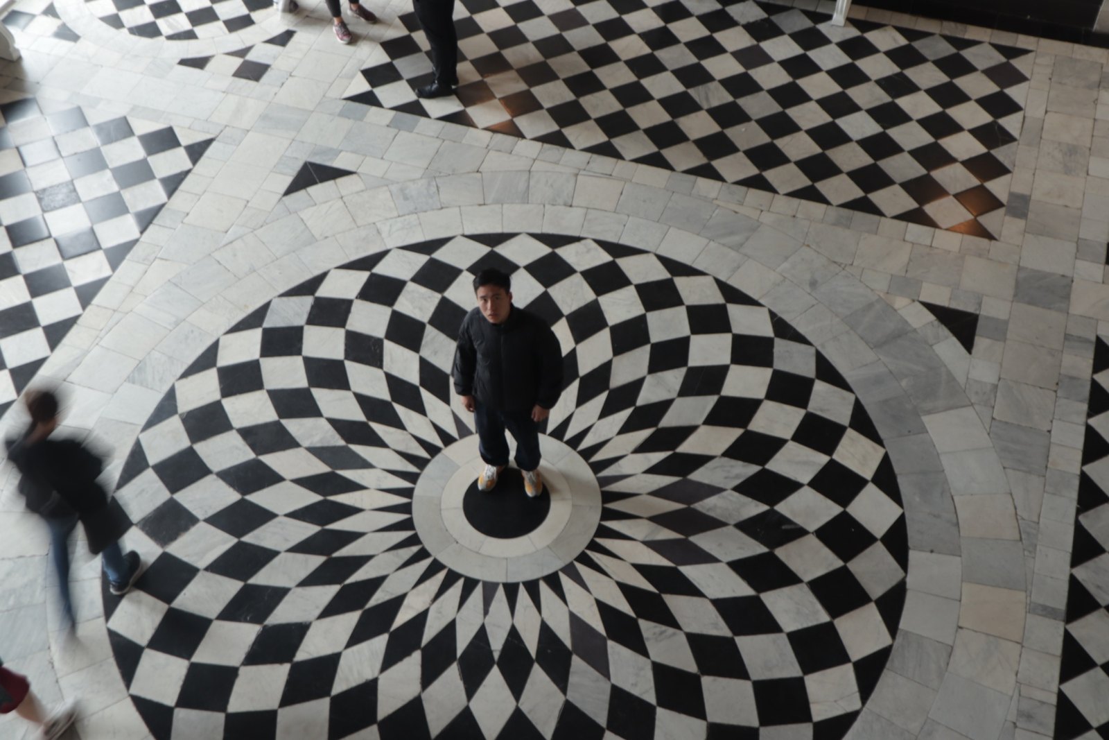 A figure stands at the centre of a vast geometric black and white marble floor, photographed from above