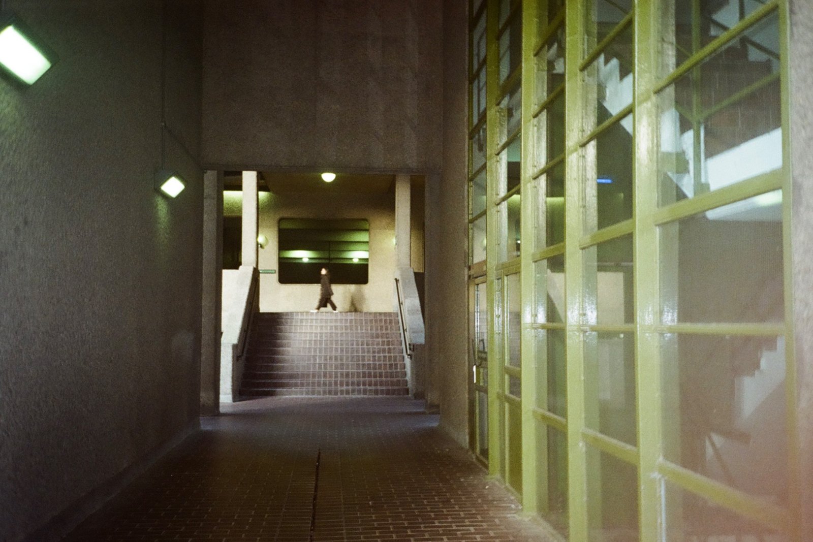 A film photograph of a brutalist corridor at night with a lone figure ascending stairs in the distance
