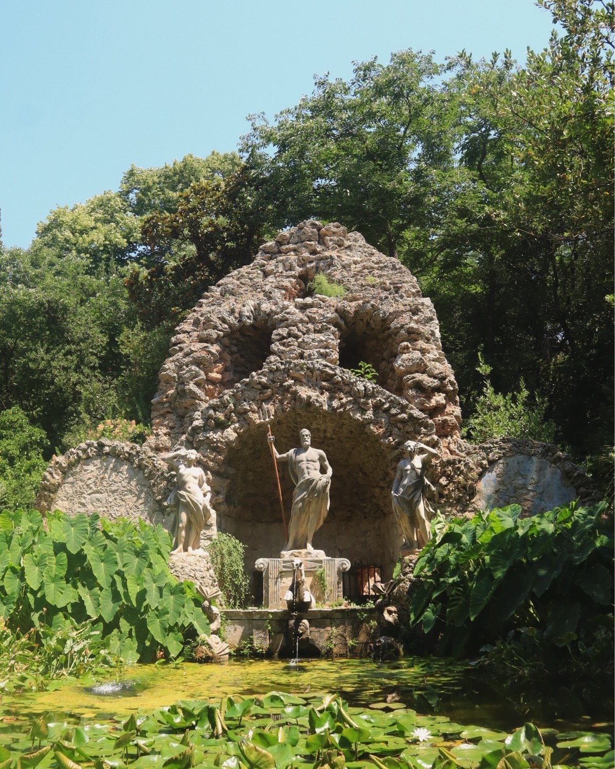 Classical statues and a Neptune fountain surrounded by lily pads and lush vegetation